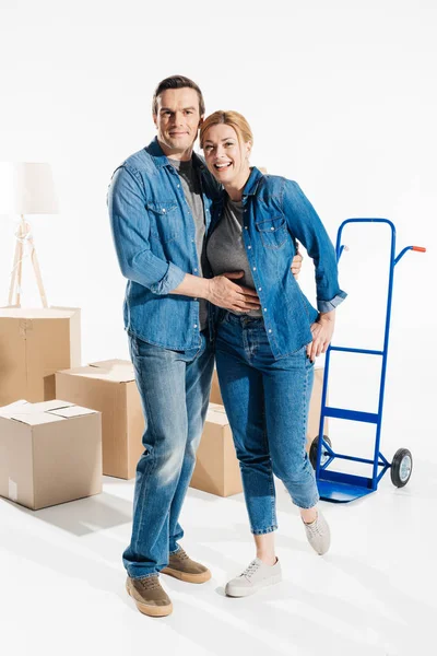 Happy couple relocating and embracing by trolley cart and boxes isolated on white — Stock Photo