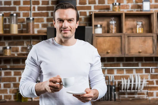 Hombre guapo sosteniendo taza de café y sonriendo a la cámara en la cocina - foto de stock