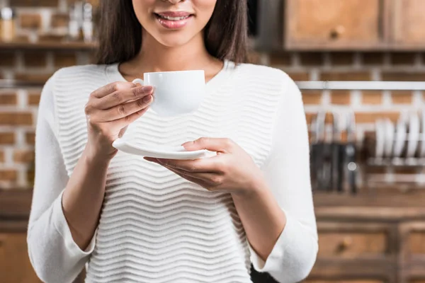 Plan recadré d'une femme afro-américaine souriante tenant une tasse de café blanche et une soucoupe — Photo de stock