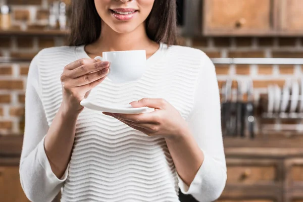 Tiro recortado de mulher americana africana sorridente segurando xícara de café branco e pires — Fotografia de Stock