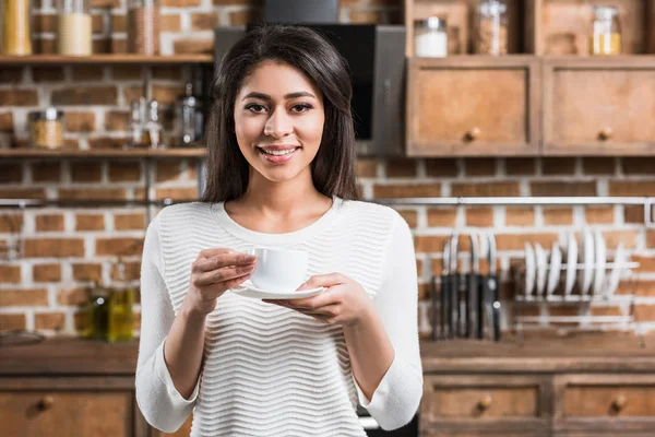 Bela menina afro-americana segurando xícara de café e sorrindo para a câmera na cozinha — Fotografia de Stock