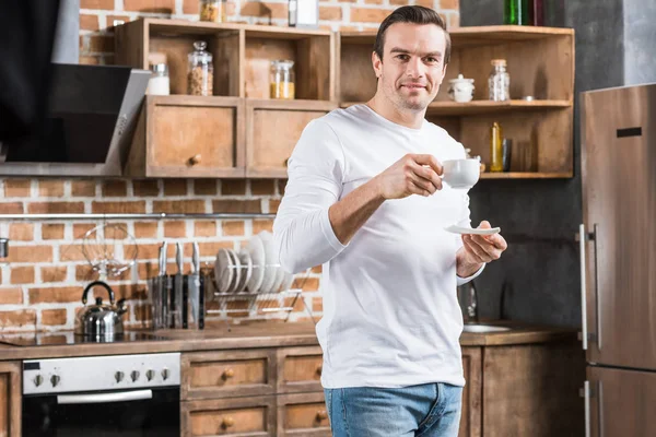 Hombre guapo sosteniendo taza de café y sonriendo a la cámara en la cocina - foto de stock