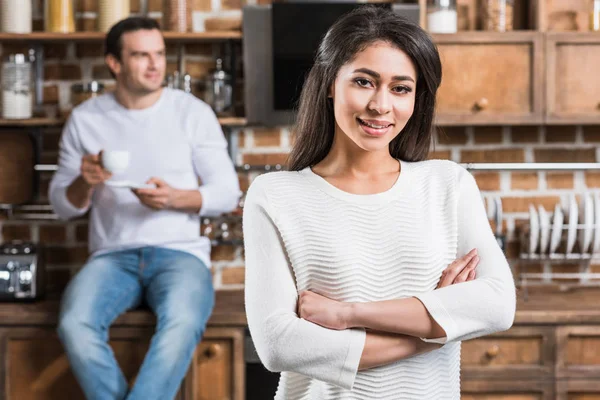 Hermosa joven afroamericana mujer de pie con los brazos cruzados y sonriendo a la cámara mientras su novio beber café detrás en la cocina - foto de stock