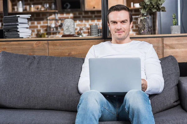 Homem bonito usando laptop e sorrindo para a câmera enquanto sentado no sofá em casa — Fotografia de Stock