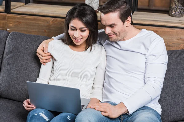 Sonriente pareja multiétnica utilizando el ordenador portátil juntos mientras se sienta en el sofá en casa - foto de stock