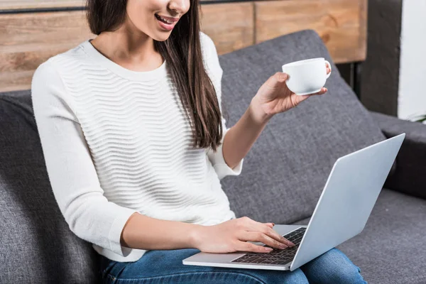 Tiro recortado de sorrir menina americana africana usando laptop e beber café no sofá — Fotografia de Stock