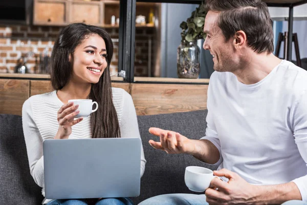 Многонациональная пара улыбается друг другу и разговаривает во время питья кофе и использования ноутбука на дому — стоковое фото
