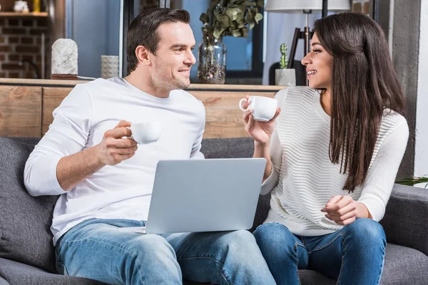 Pareja multiétnica sosteniendo tazas de café y sonriendo entre sí mientras se utiliza el ordenador portátil en casa - foto de stock