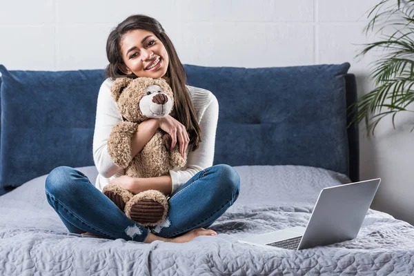 Красива щаслива афроамериканська дівчина обіймає плюшевого ведмедя і посміхається на камеру, сидячи на ліжку з ноутбуком — стокове фото