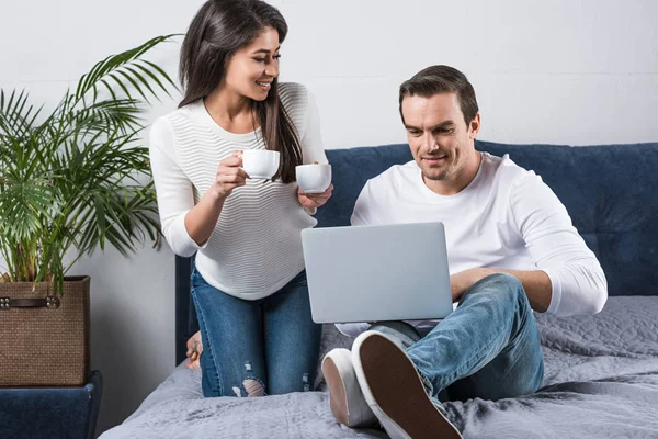 Sorrindo mulher afro-americana segurando xícaras de café e olhando para o namorado usando laptop na cama — Fotografia de Stock