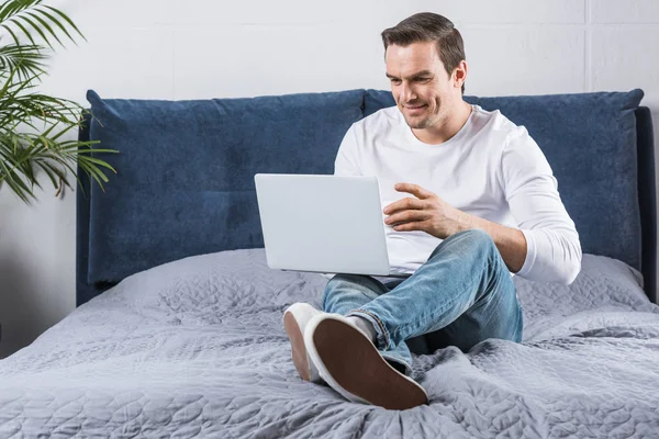 Bel homme souriant utilisant un ordinateur portable sur le lit — Photo de stock