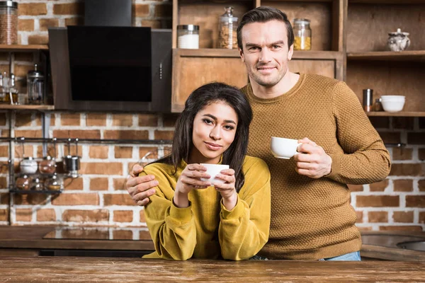 Glückliches multiethnisches Paar, das Kaffee trinkt und in der Küche in die Kamera lächelt — Stockfoto