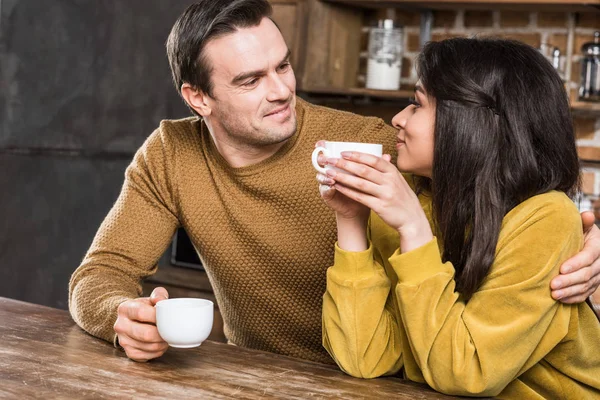 Glückliches multiethnisches Paar trinkt Kaffee und lächelt sich zu Hause an — Stockfoto