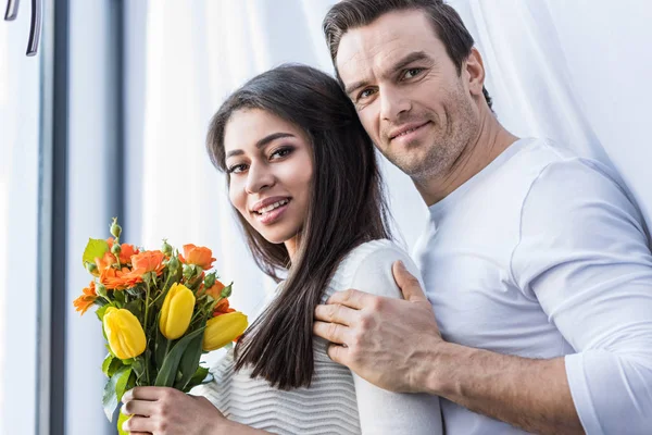 Glückliches multiethnisches Paar, das sich umarmt und in die Kamera lächelt, während Frau einen schönen Blumenstrauß in der Hand hält — Stockfoto