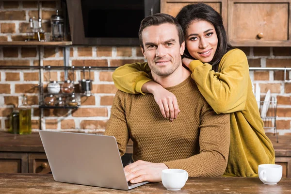 Glückliches junges multiethnisches Paar lächelt in die Kamera, während Mann Laptop benutzt — Stockfoto