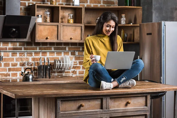 Sorrindo menina americana africana segurando xícara de café e usando laptop enquanto sentado no topo da mesa de madeira na cozinha — Fotografia de Stock