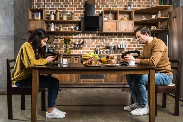 Vista laterale della giovane coppia multietnica che utilizza gli smartphone durante la colazione — Foto stock