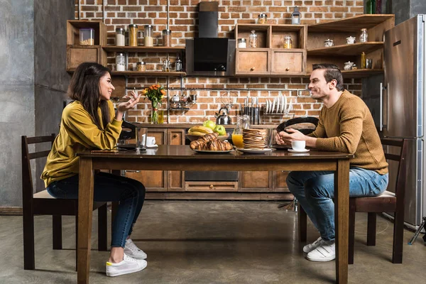 Vista lateral do jovem casal multiétnico sorrindo uns aos outros e conversando durante o café da manhã — Fotografia de Stock