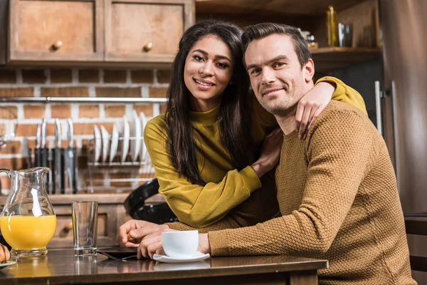 Felice giovane coppia multietnica sorridente alla macchina fotografica mentre fa colazione a casa — Foto stock