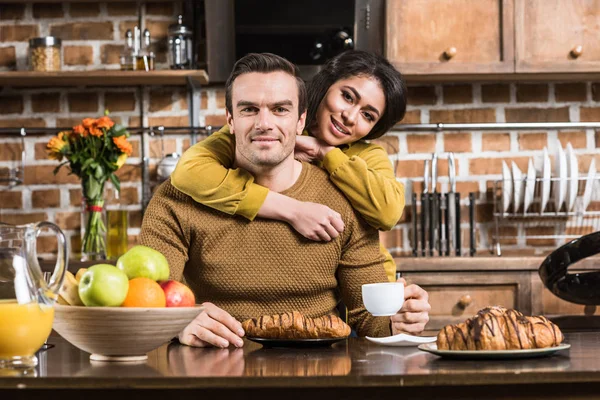 Heureux jeune couple multiethnique embrasser et sourire à la caméra tout en prenant le petit déjeuner ensemble à la maison — Photo de stock