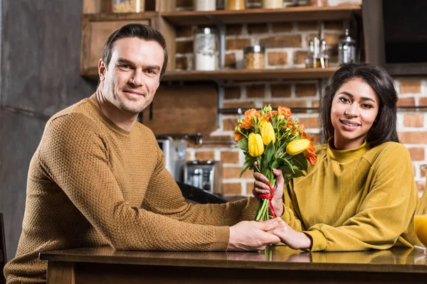 Heureux jeune couple multiethnique avec bouquet de fleurs tenant la main et souriant à la caméra à la maison — Photo de stock