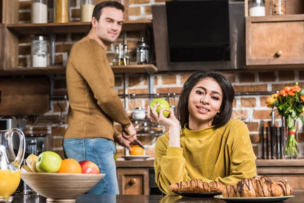 Hermosa joven afroamericana mujer sosteniendo manzana y sonriendo a la cámara mientras marido preparando el desayuno detrás - foto de stock