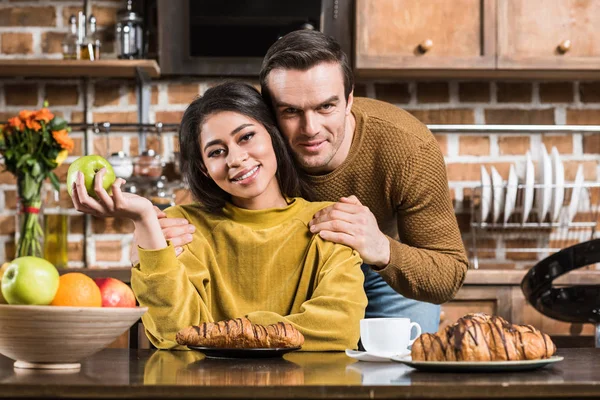 Glückliches junges multiethnisches Paar, das beim gemeinsamen Frühstück zu Hause in die Kamera lächelt — Stockfoto