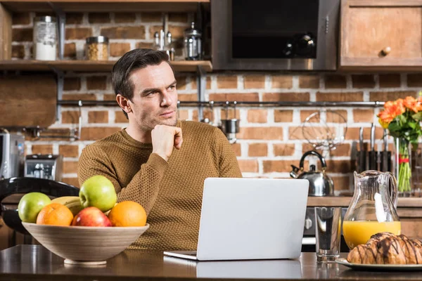 Nachdenklicher Mann schaut weg, während er Laptop am Küchentisch benutzt — Stockfoto