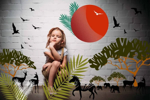 Enfant femelle en costume de safari et chapeau assis au mur blanc avec illustration d'animaux safari — Photo de stock