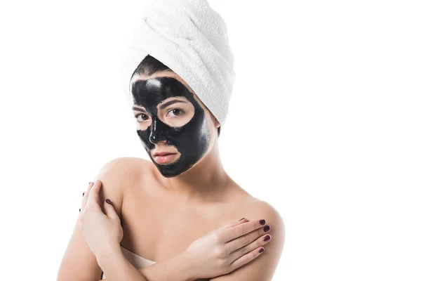Hermosa chica en cosmética máscara facial de arcilla negra y toalla en la cabeza mirando a la cámara aislada en blanco - foto de stock