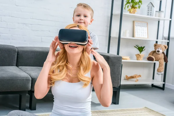 Mãe usando fones de ouvido de realidade virtual com o pequeno filho parado atrás — Fotografia de Stock