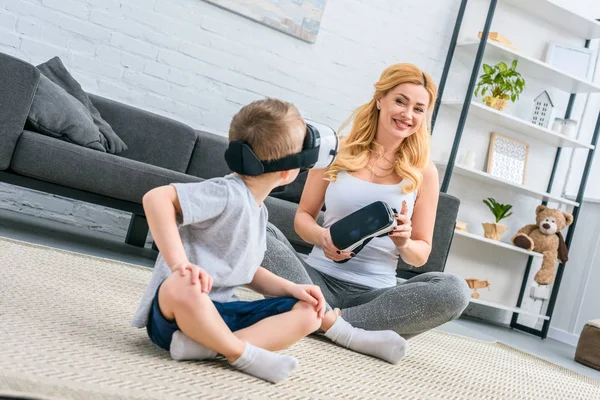 Niño pequeño usando auriculares de realidad virtual y sentado con la madre - foto de stock