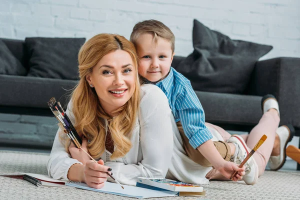 Happy little son sitting on mother back while she painting at home — Stock Photo