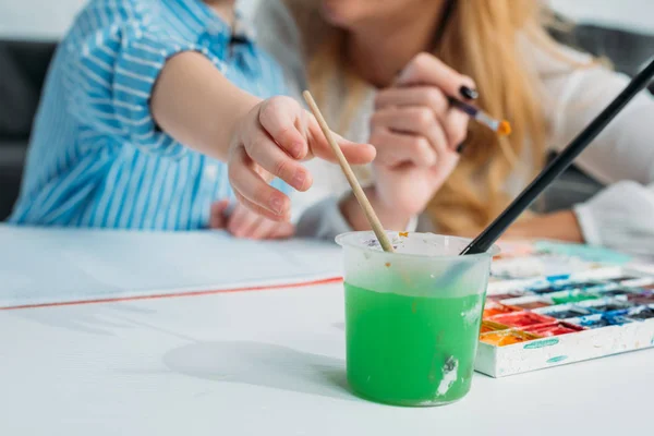 Cropped image of mother and son dipping paintbrush into colored water — Stock Photo