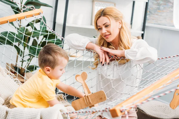 Мама смотрит, как маленький мальчик играет с деревянной игрушкой самолета — стоковое фото