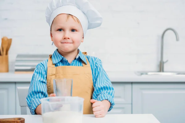 Garçon en chapeau de cuisine et tablier tenant le verre dans la cuisine — Photo de stock