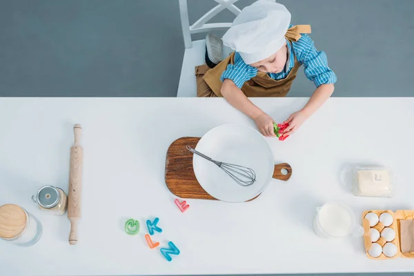 Top view of little boy in cooking hat sitting at table with kitchenware and ingredients — Stock Photo