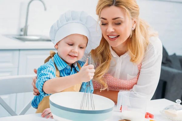 Mère souriante embrassant petit fils alors qu'il utilise batteur à main — Photo de stock