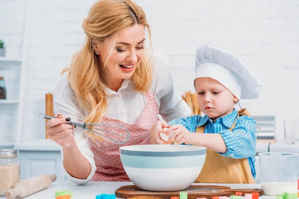 Madre sorridente con battitore a mano e bambino che versa l'uovo nella ciotola — Foto stock