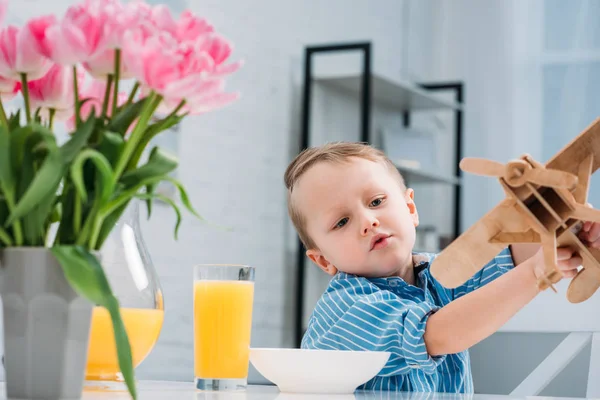 Little boy playing with wooden airplane at table with plate, juice and tulips — Stock Photo