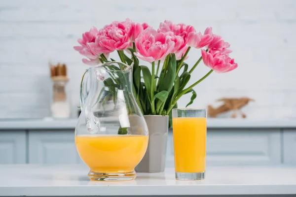 Front view of orange juice and vase with pink tulips on table in modern kitchen — Stock Photo