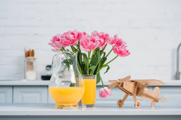 Rosa Tulpen in der Vase, Orangensaft und Holzflugzeug auf dem Tisch in der modernen Küche — Stockfoto