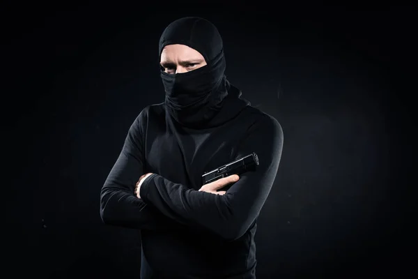 Thief in black balaclava holding gun in folded arms on black — Stock Photo