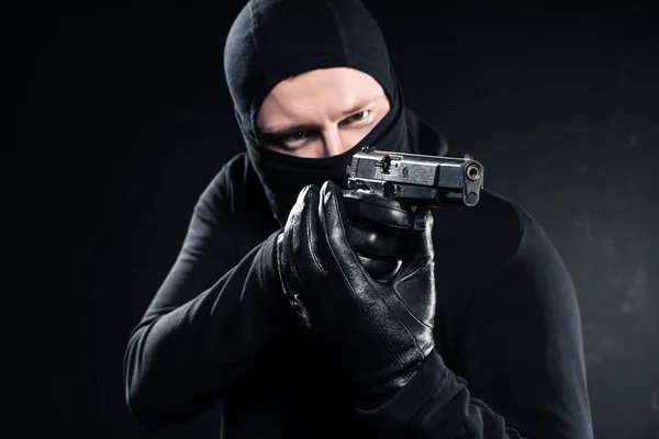 Male robber in balaclava aiming with gun on black — Stock Photo