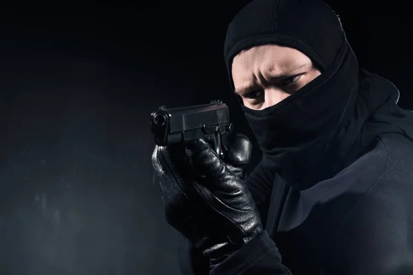 Robber in balaclava and gloves aiming with gun on black — Stock Photo