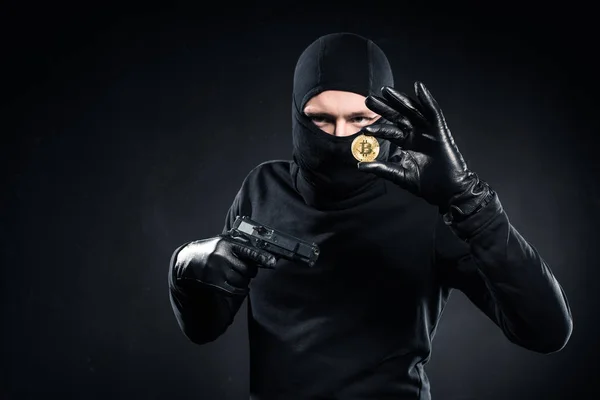 Man in black balaclava holding gun and golden bitcoin — Stock Photo