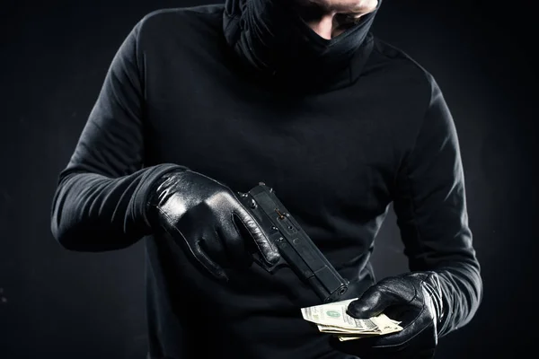 Man in black balaclava holding gun and dollars — Stock Photo