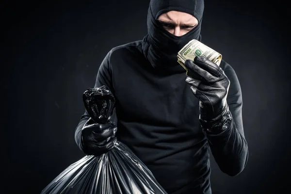 Man in balaclava holding plastic bag with cash and dollars — Stock Photo