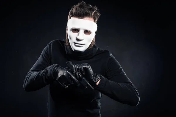 Ladrón con máscara blanca y ropa negra recargando su arma - foto de stock