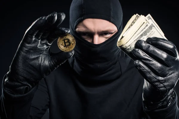 Hombre criminal en pasamontañas sosteniendo dólares y bitcoin - foto de stock